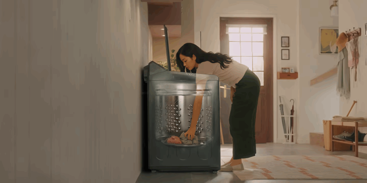 Máy giặt cửa trên màu xám với lồng giặt trong suốt, người phụ nữ đang cho quần áo vào máy